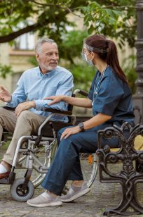 Full length shot of cheerful mature man, recovering patient in wheelchair talking to caring nurse, resting in the park near hospital. Medical insurance, rehabilitation concept